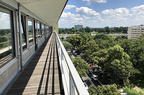 Vue depuis un balcon le long d'un long corridor avec des balustrades sur la gauche. Vaste vue sur la cime d'arbres verts, des structures urbaines et des voitures garées. Au loin, un autre bâtiment et un plan d'eau qui reflète la lumière du soleil. Ciel partiellement nuageux avec la lumière du soleil qui projette des ombres sur le sol du balcon.